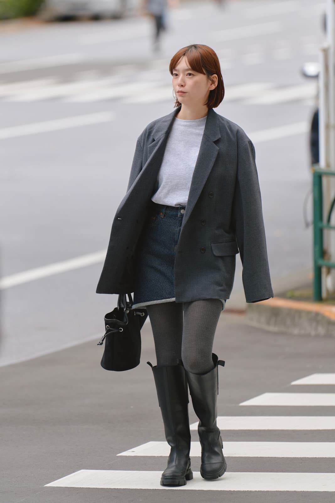 Woman walking on a crosswalk wearing a gray oversized blazer, denim skirt, gray tights, and black knee-high boots, carrying a black handbag.
