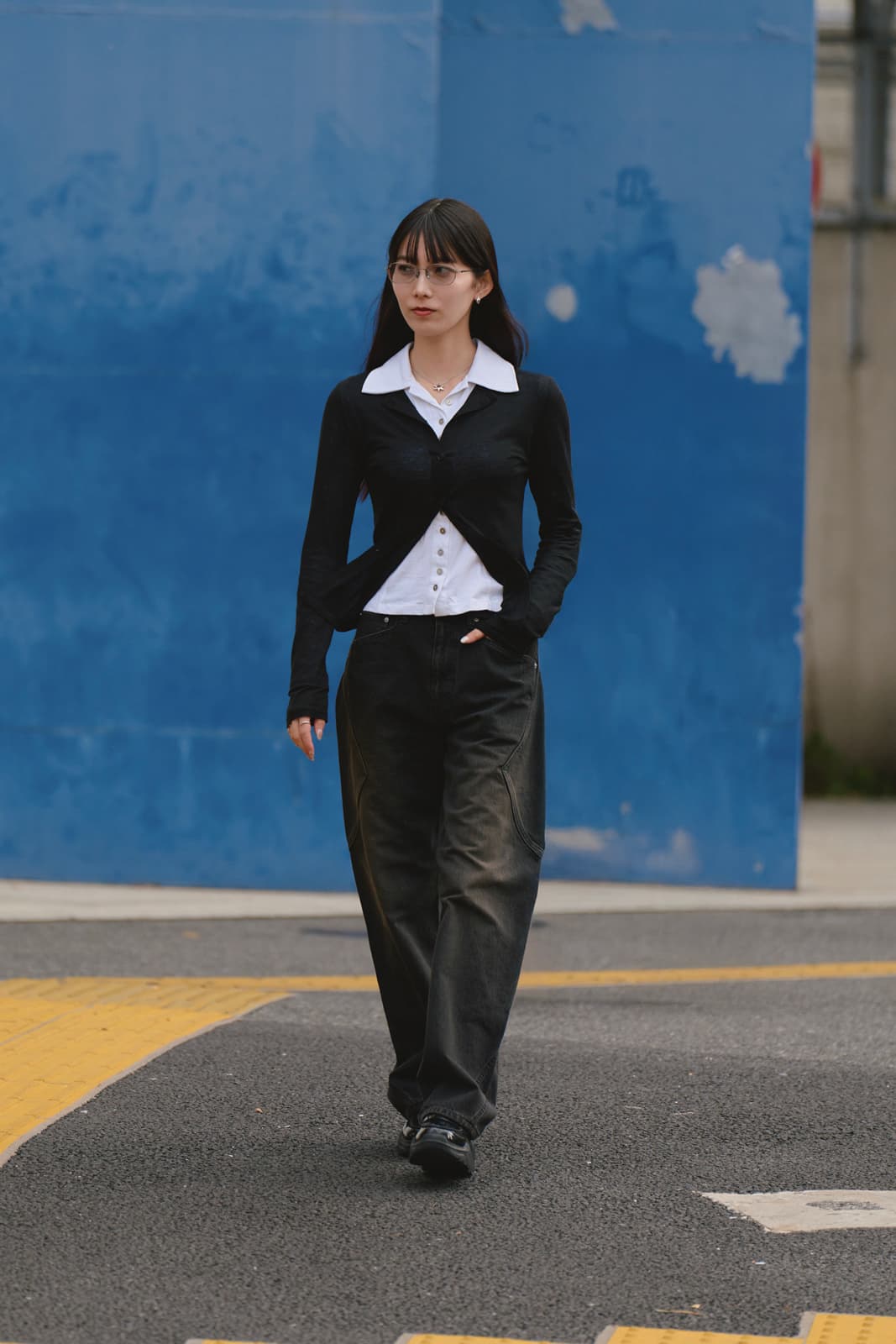 Woman with long dark hair, glasses, black cardigan, white collared shirt, and loose black pants walking on asphalt with blue wall background.