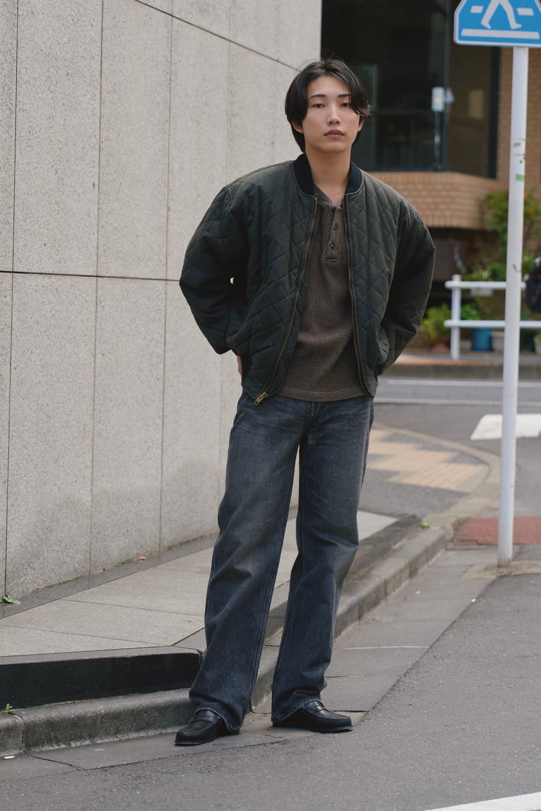Young man standing on sidewalk with hands in the pockets of a dark quilted jacket, wearing loose dark jeans and black shoes.