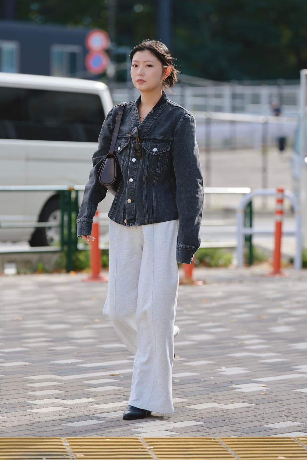 Woman walking outdoors wearing a black denim jacket, loose white pants, black shoes, and carrying a brown shoulder bag.