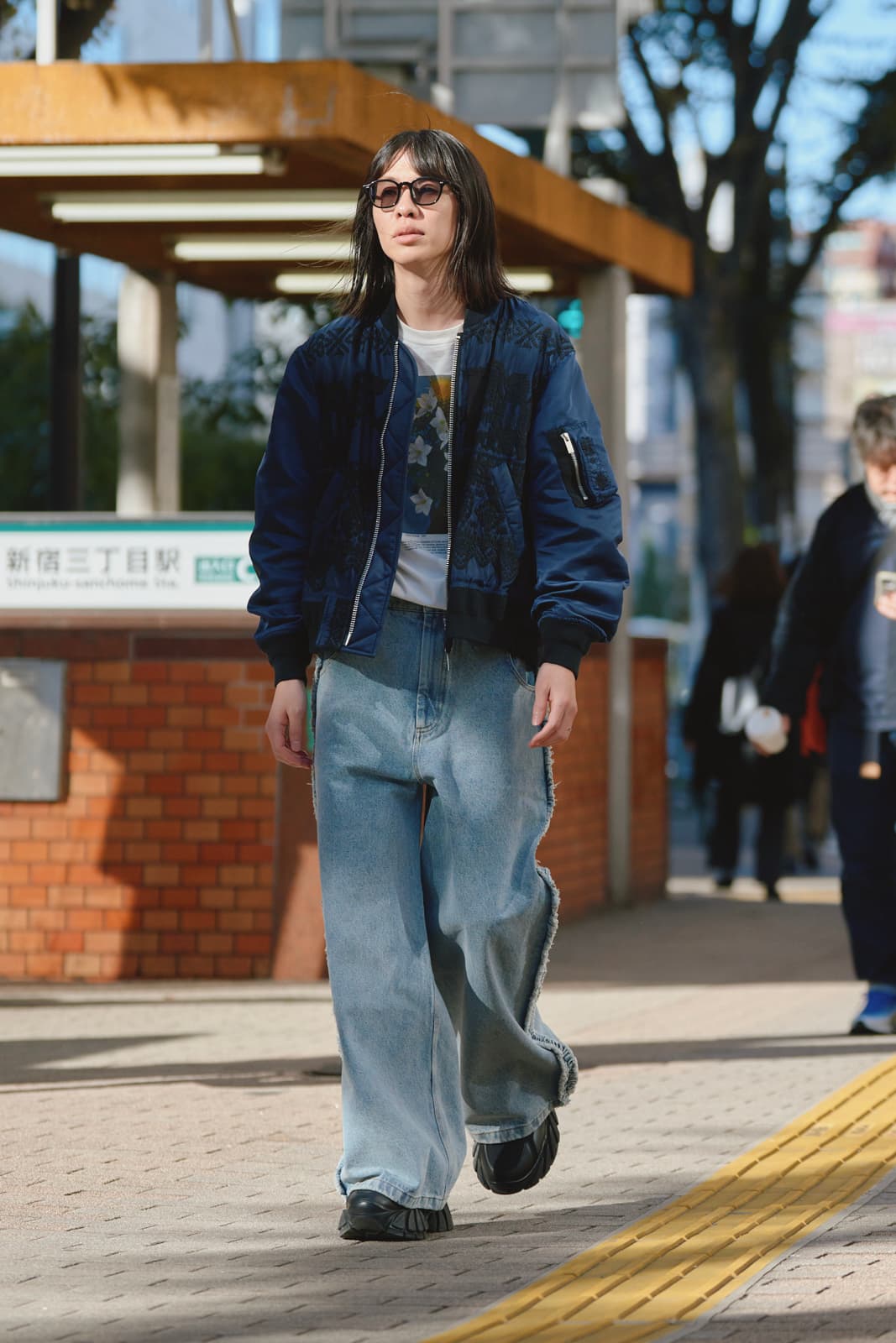 Person wearing sunglasses, a patterned navy bomber jacket, white graphic t-shirt, and wide light blue jeans walking on a city sidewalk.
