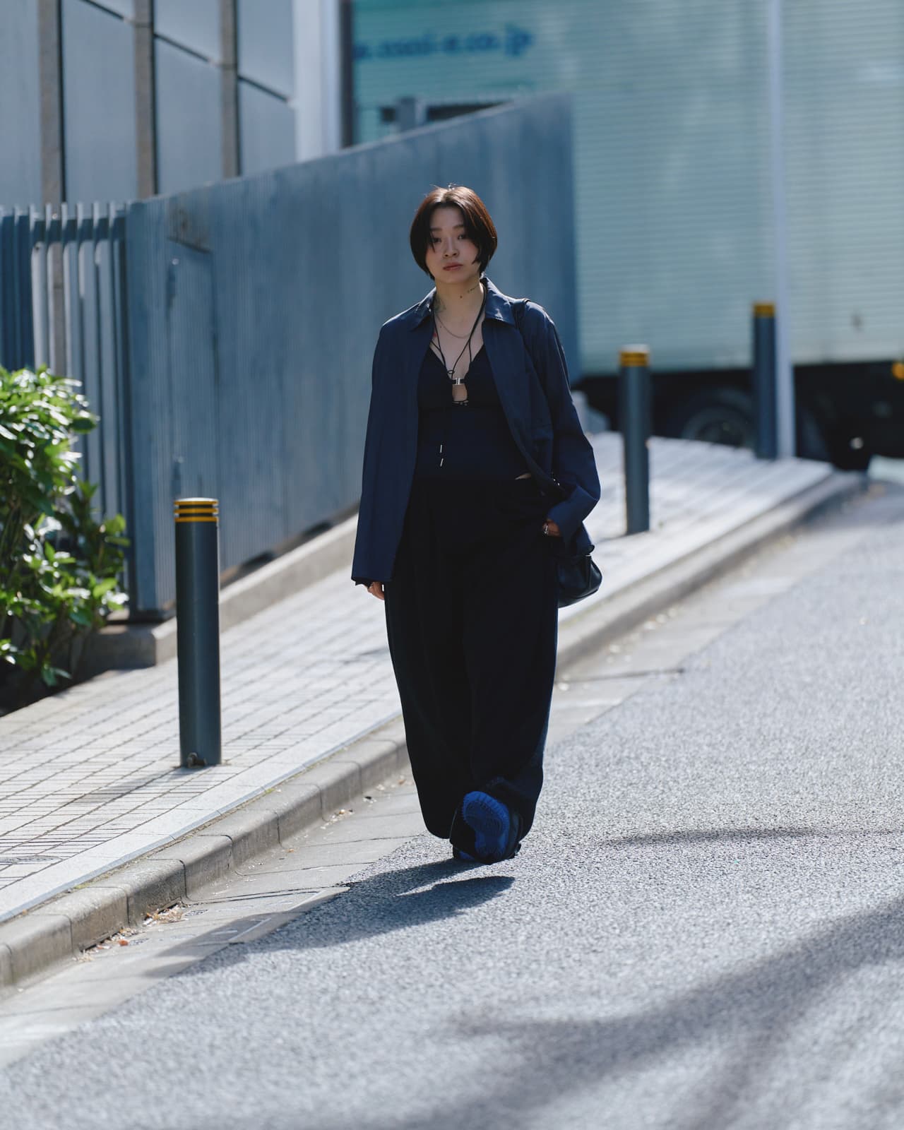Person walking down a sunny street wearing a dark loose-fitting outfit with a jacket and necklace.