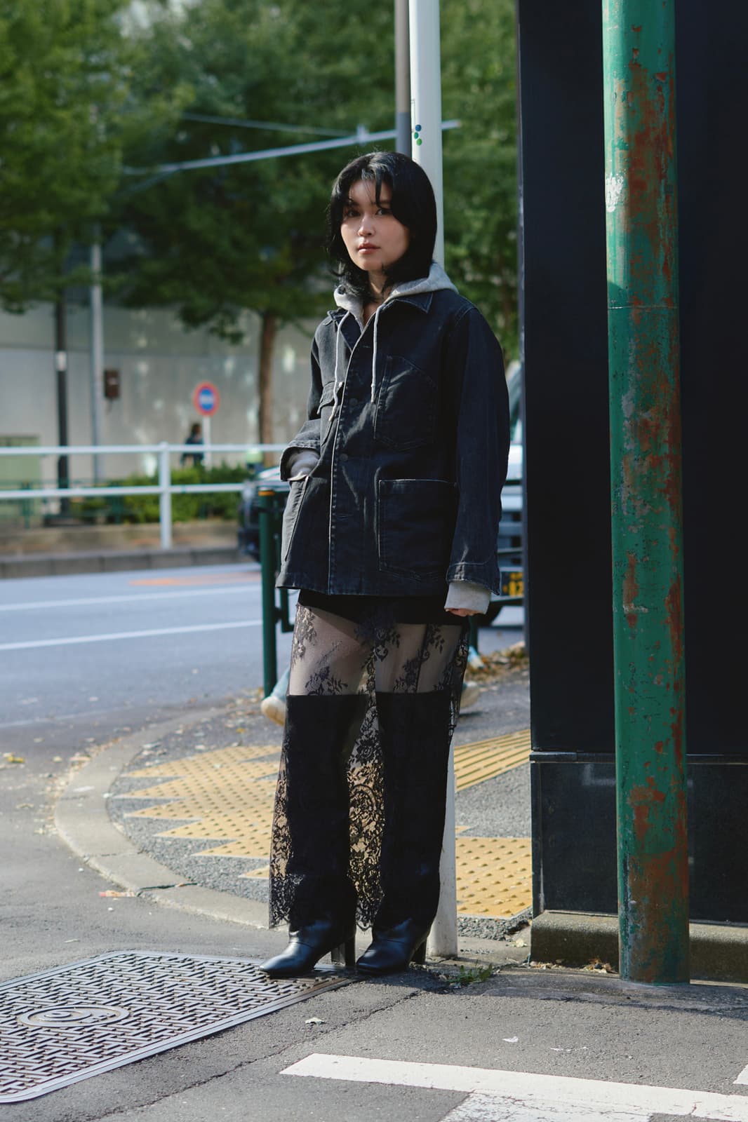 Person standing on a street corner wearing a dark jacket, grey hoodie, black lace skirt, and black heeled boots.
