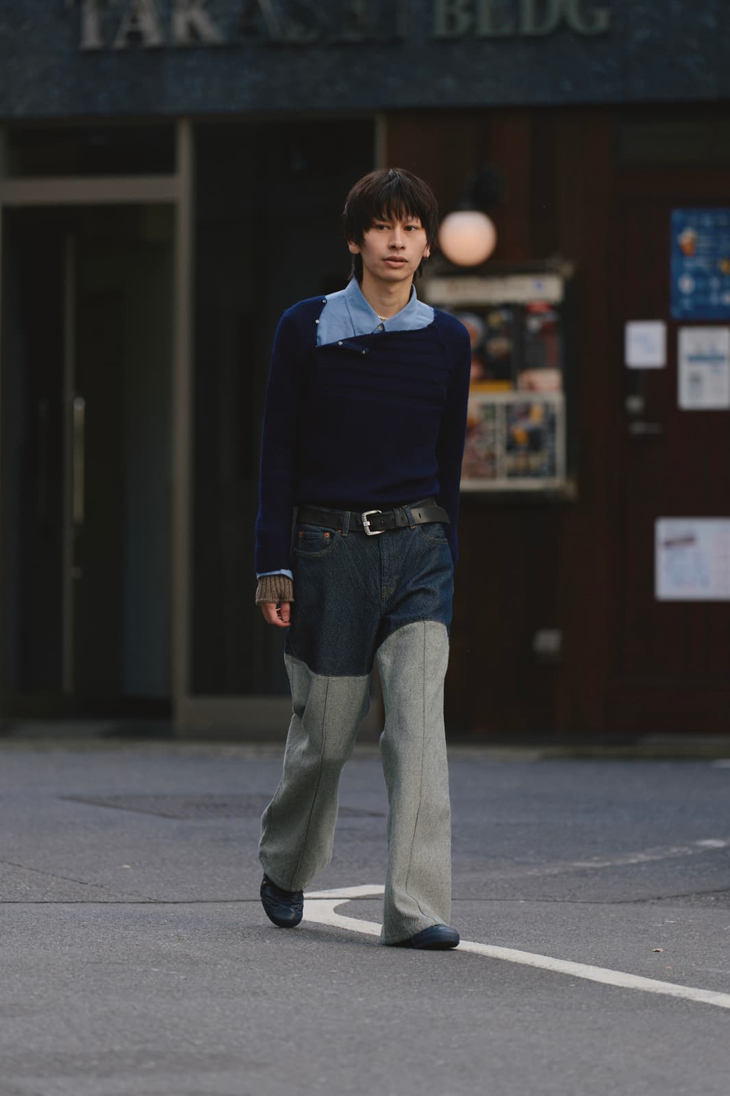 Young person walking on a street wearing a navy sweater over a blue collared shirt and two-tone wide-leg pants.