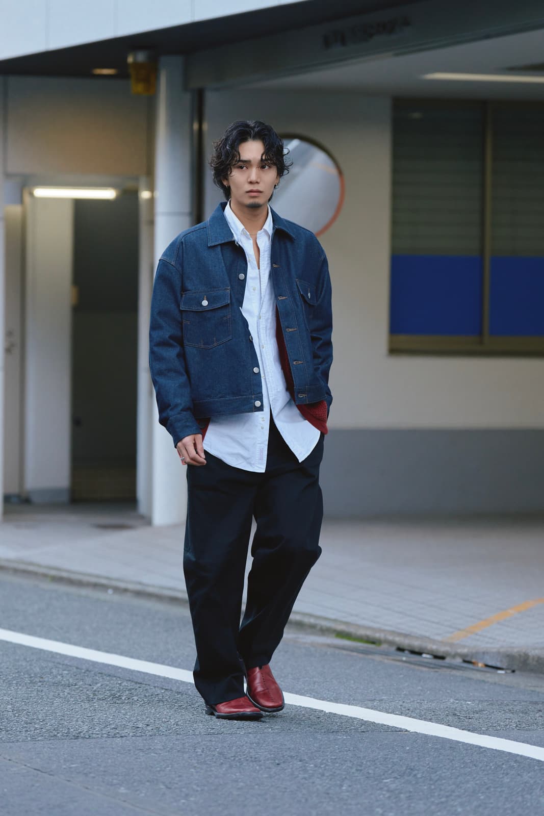Man with wavy black hair wearing a denim jacket, white shirt, black pants, and red shoes walking on a street.