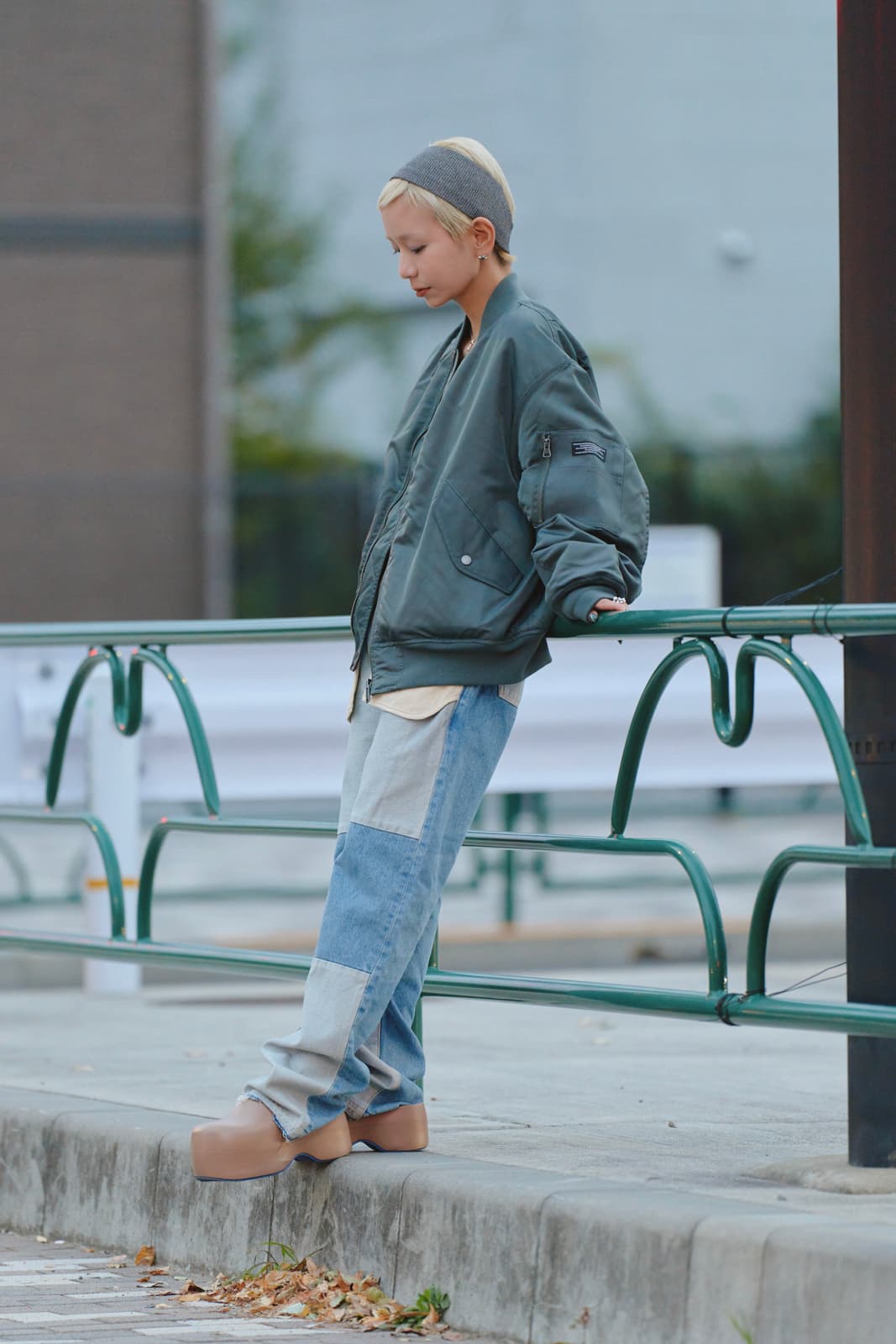 Person with short blonde hair wearing a gray headband, oversized green jacket, patchwork blue jeans, and tan platform shoes leaning against a green metal railing on a sidewalk.