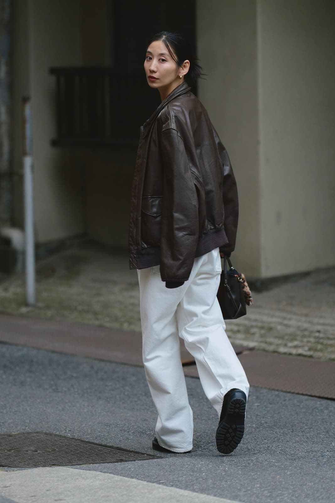 Woman turning back walking on street wearing a dark brown leather jacket, white pants, and black shoes while carrying a black handbag.