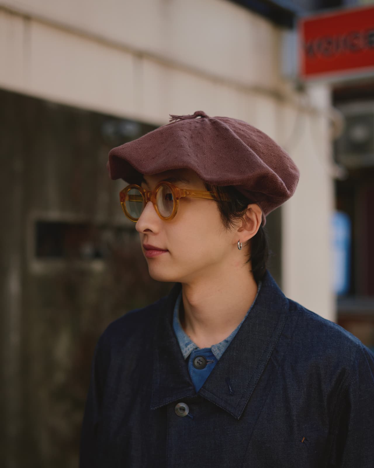 Young person in profile wearing a brown beret, round amber glasses, a small hoop earring, and a dark jacket.