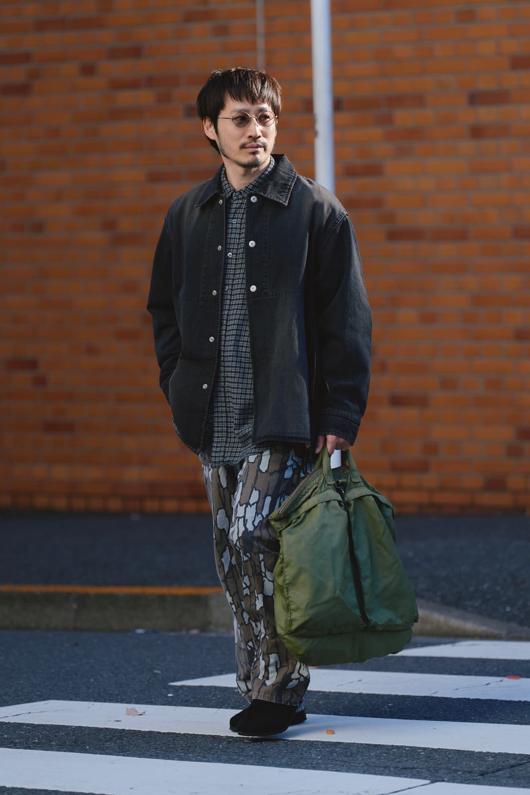 Man wearing sunglasses, a black jacket, patterned pants, and carrying a green bag walking on a zebra crossing.