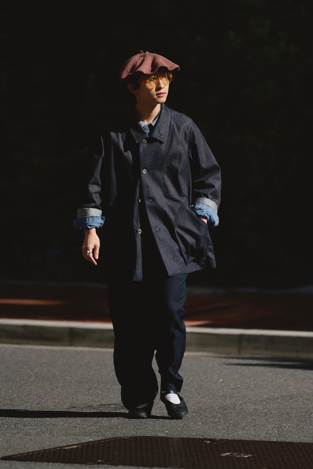 Person wearing a brown beret, amber glasses, dark oversized jacket with rolled-up sleeves, and black pants walking on a street.