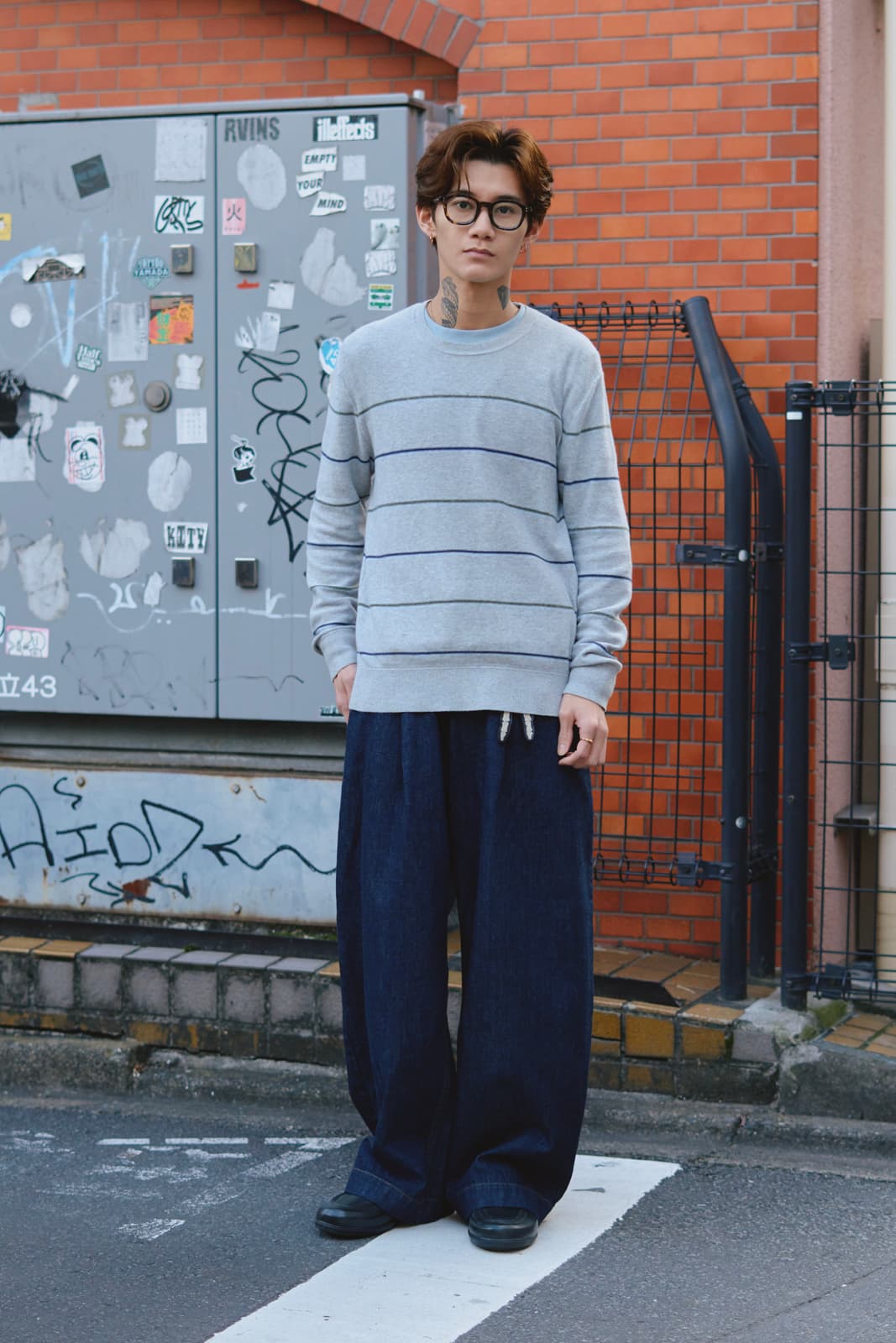 Young person with glasses and neck tattoos wearing a gray striped sweater and wide navy pants standing on a street crosswalk.